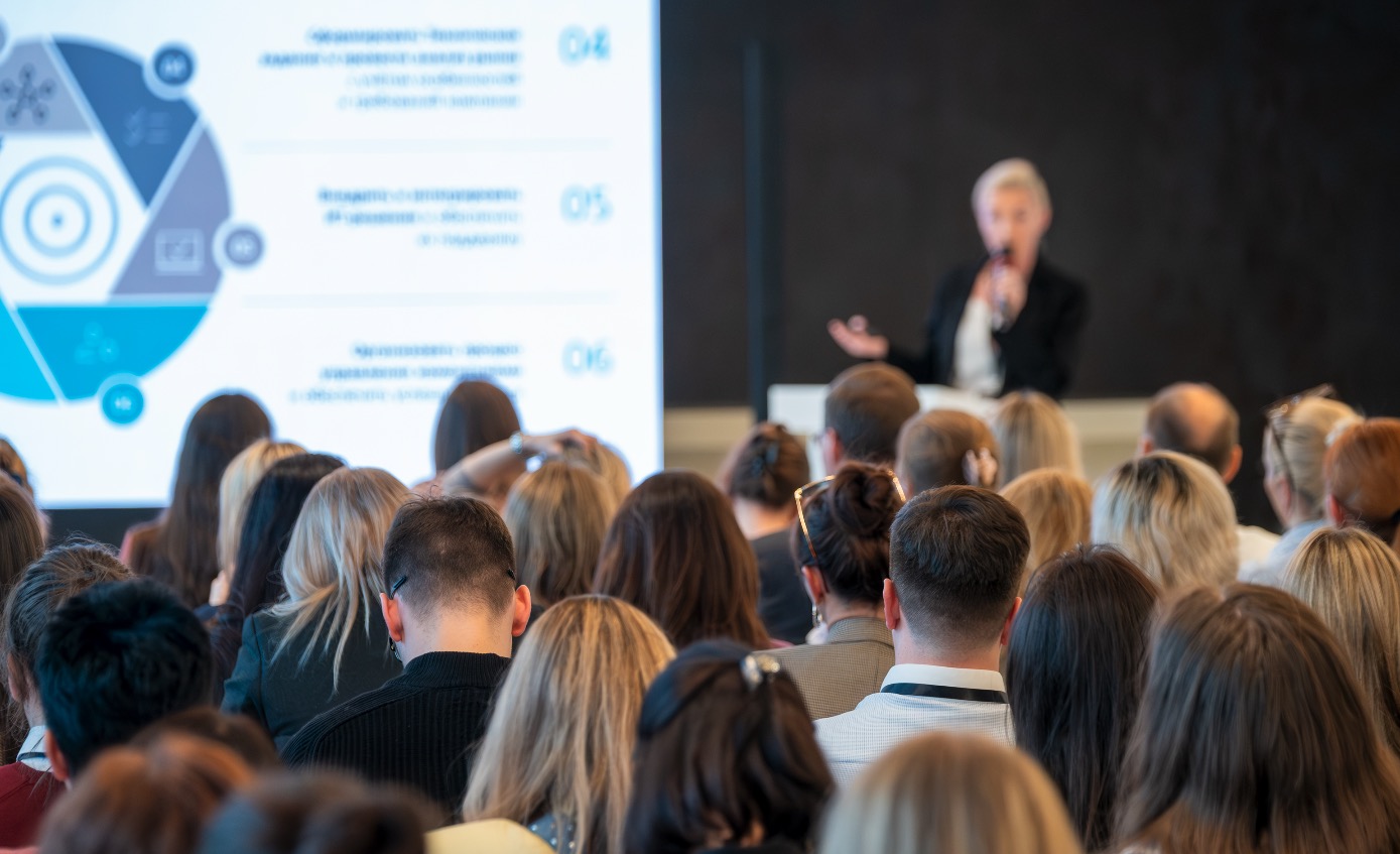 A corporate presenter addressing a large audience of professionals at a work gathering.